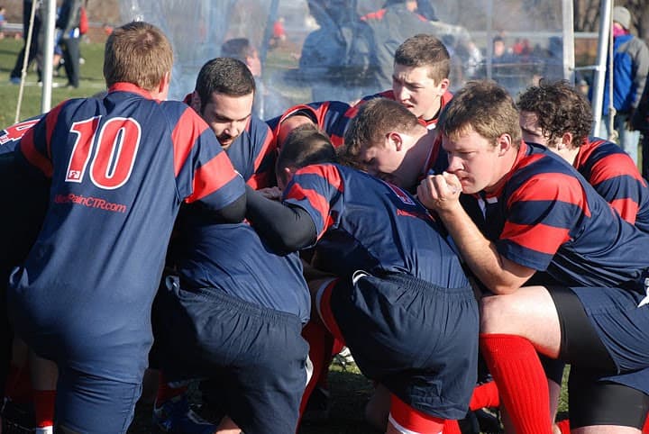 Sacred Heart Rugby forwards binding into a scrum, shoulders locked and heads down, in navy and red hooped jerseys.