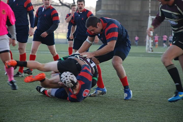 A Sacred Heart Rugby player driving low into a tackle and bringing an opposing ball carrier to ground.