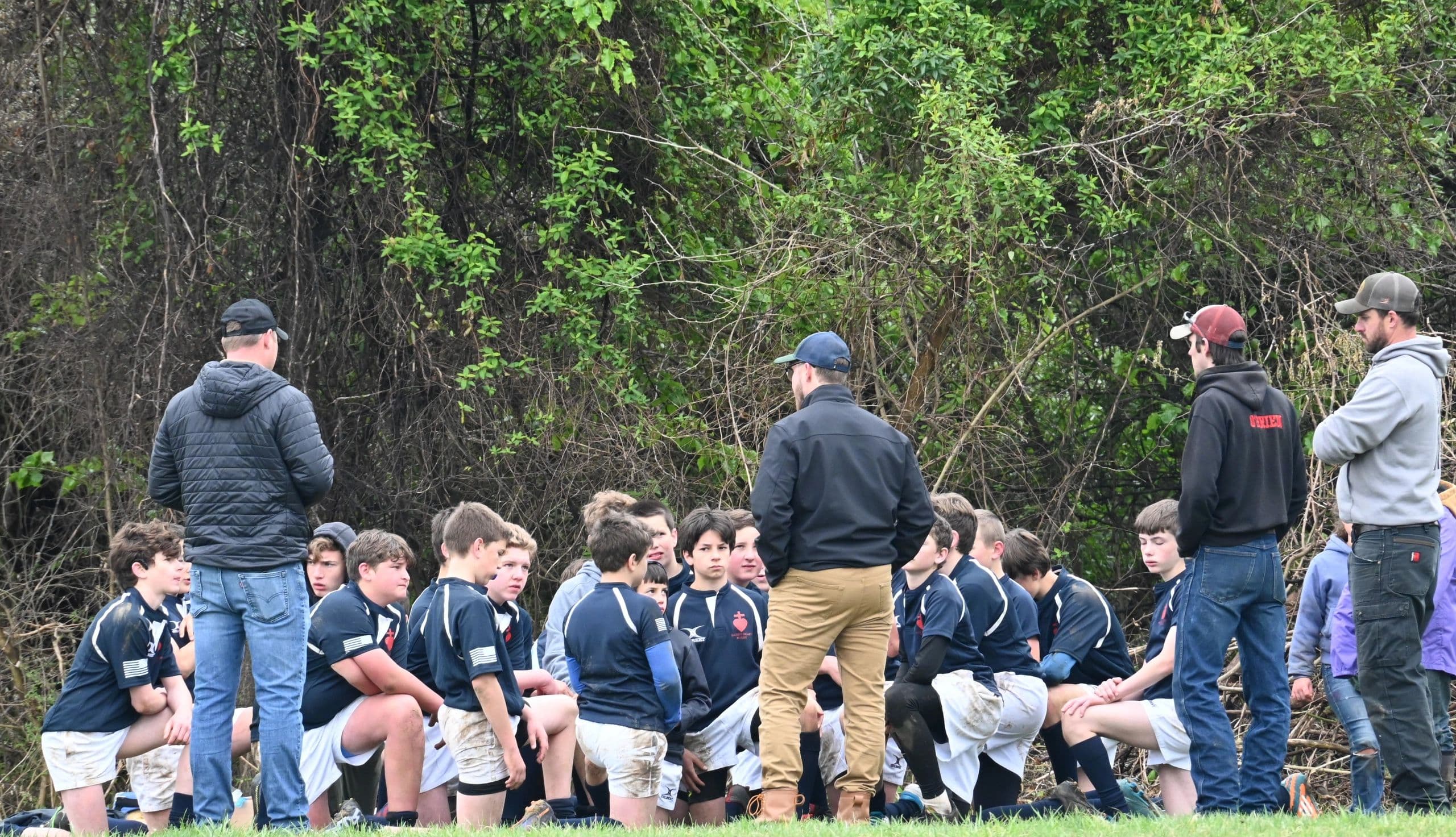 Sacred Heart Rugby players kneeling in navy jerseys while a coach addresses them beside the practice pitch.