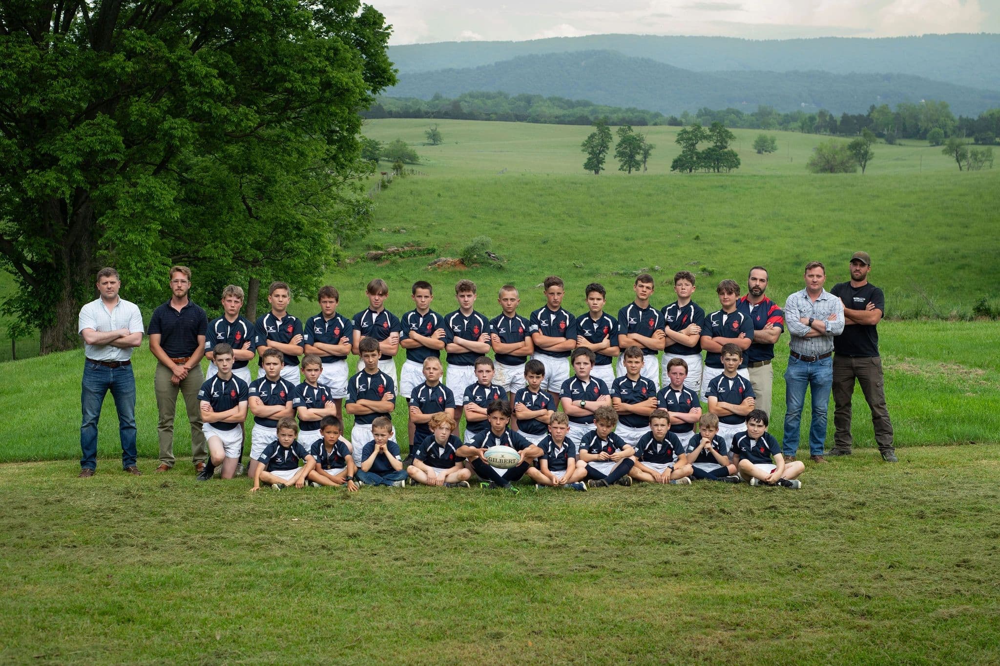 The Sacred Heart Rugby squad and coaches assembled on a grass pitch with the Blue Ridge mountains behind them.